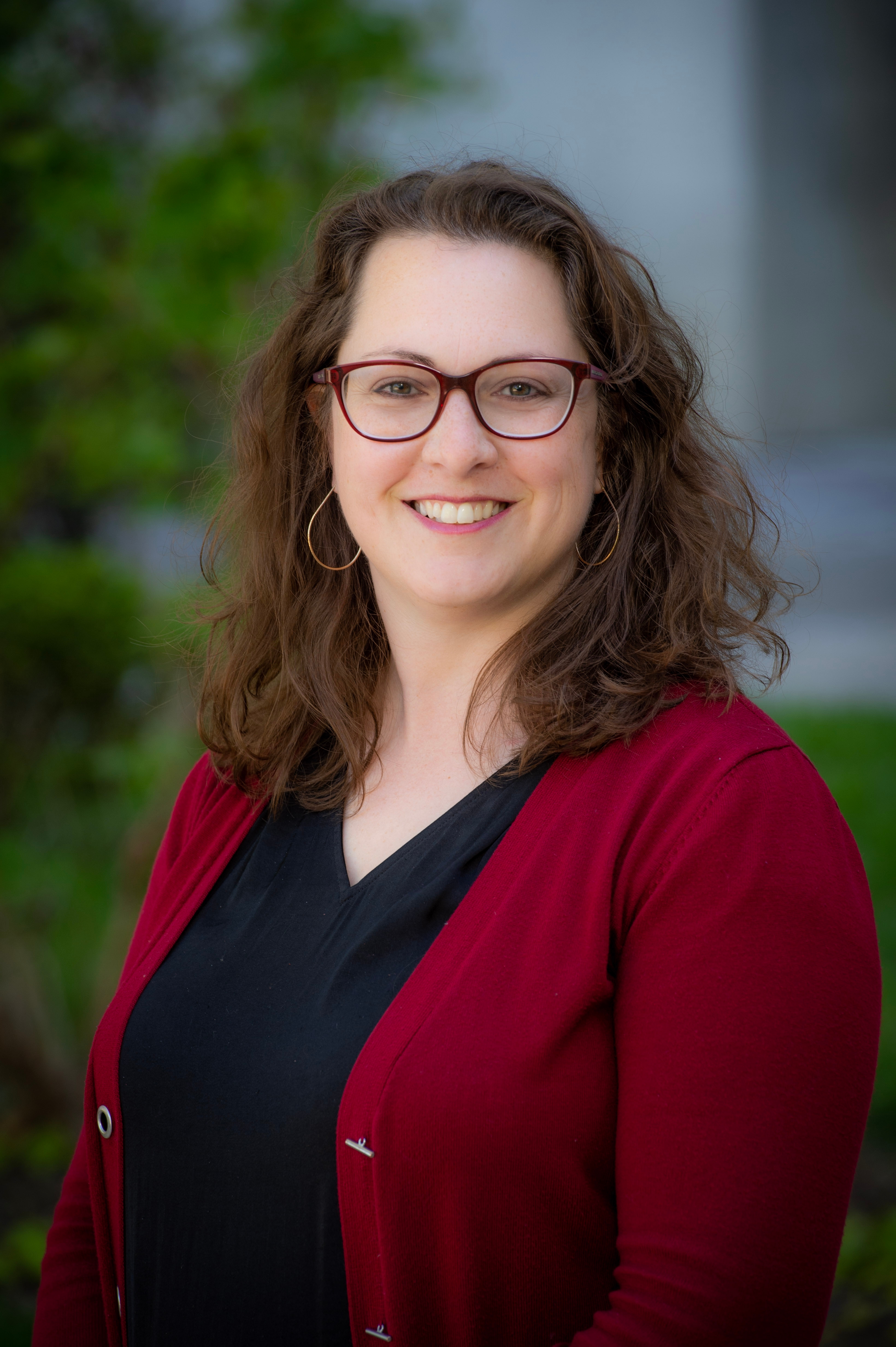 Smiling woman with wavy brown hair and red glasses wearing a burgundy cardigan and black top, posed outdoors against a blurred greenery background.