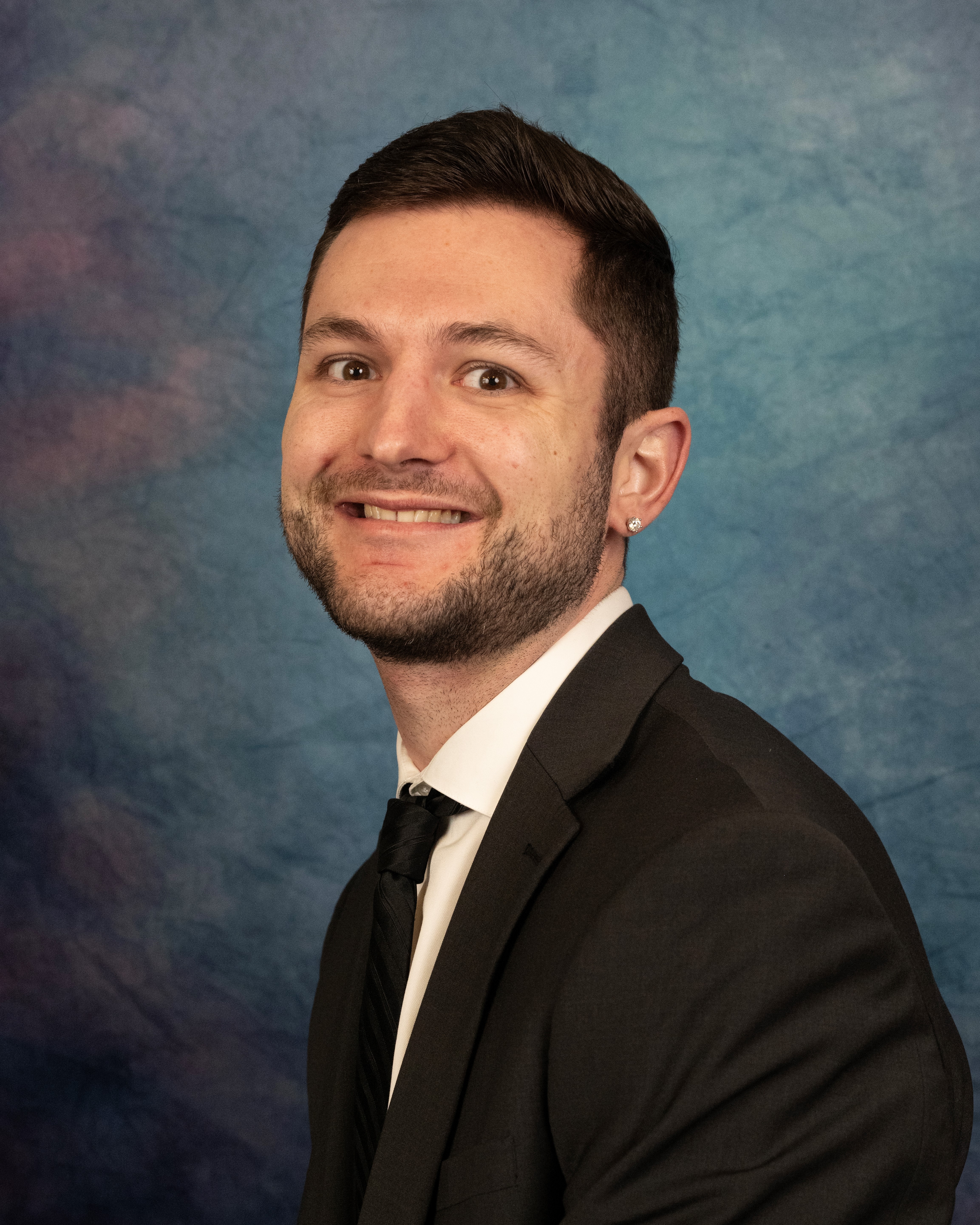 Portrait of a smiling person in a black suit and tie against a blue studio backdrop.