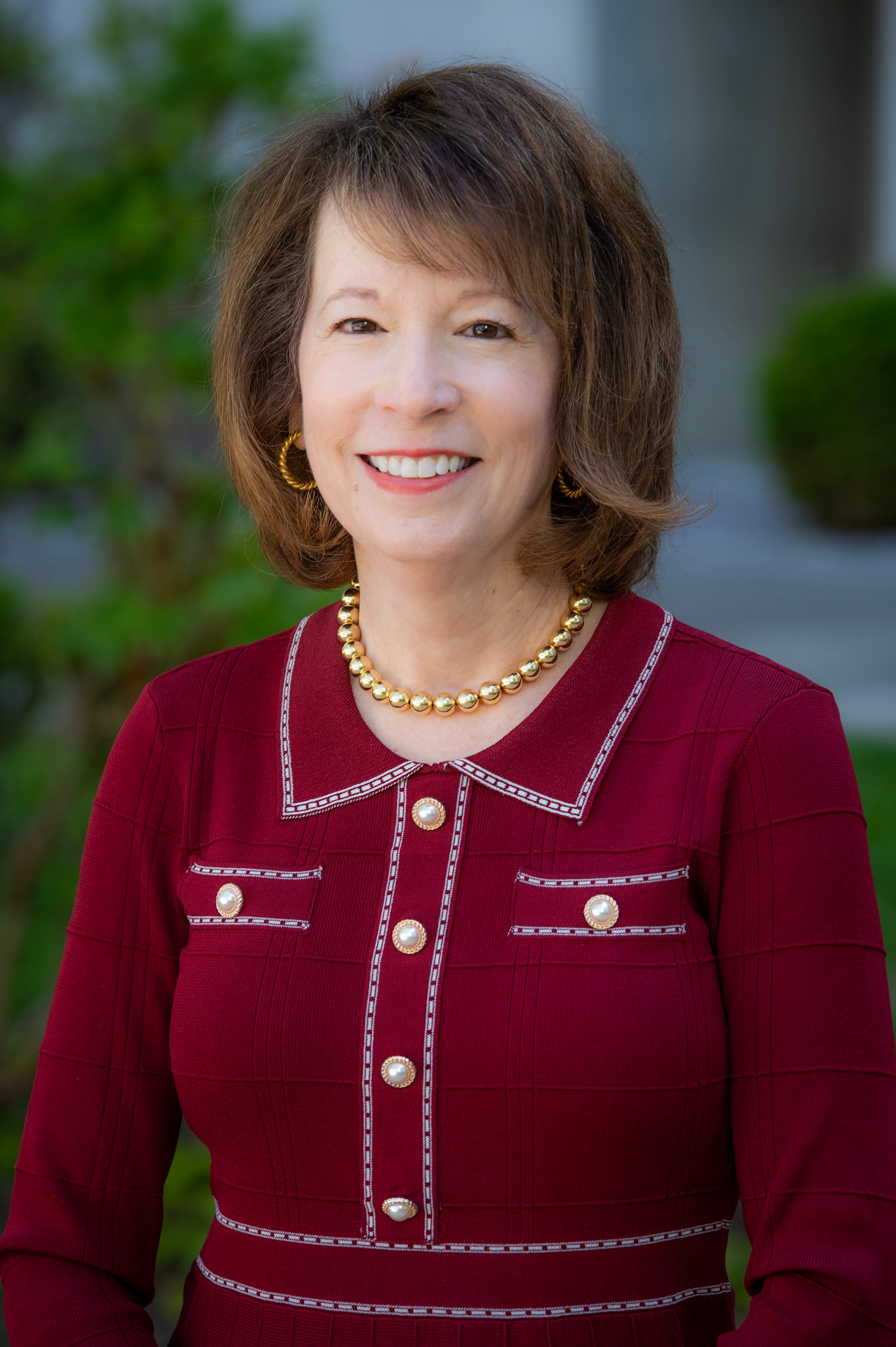 Smiling woman in a burgundy jacket with gold buttons and jewelry, posed outdoors with a softly blurred background.