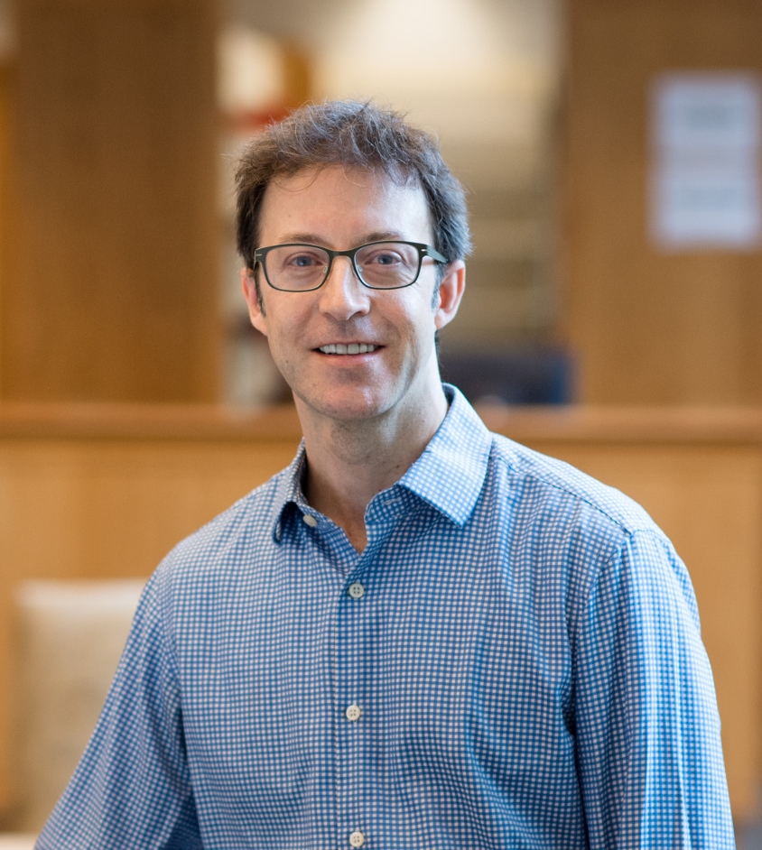 Portrait of a smiling man wearing glasses and a blue checkered button-down shirt, standing indoors with a blurred background.