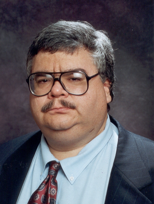 Studio portrait of a middle-aged man with gray hair, a mustache, and large glasses, wearing a suit jacket and patterned tie against a dark background.