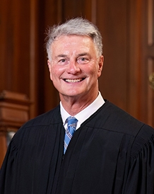 Smiling man in a black judicial robe and blue striped tie standing in a wood-paneled room.