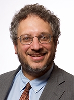 Smiling middle-aged man with curly hair and glasses, wearing a gray suit, light blue shirt, and patterned tie against a white background.