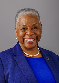 Smiling older woman with short gray hair wearing a royal blue blazer and pearl necklace against a gray studio background.