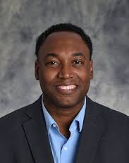 Smiling man in a dark blazer and light blue shirt against a gray studio backdrop.