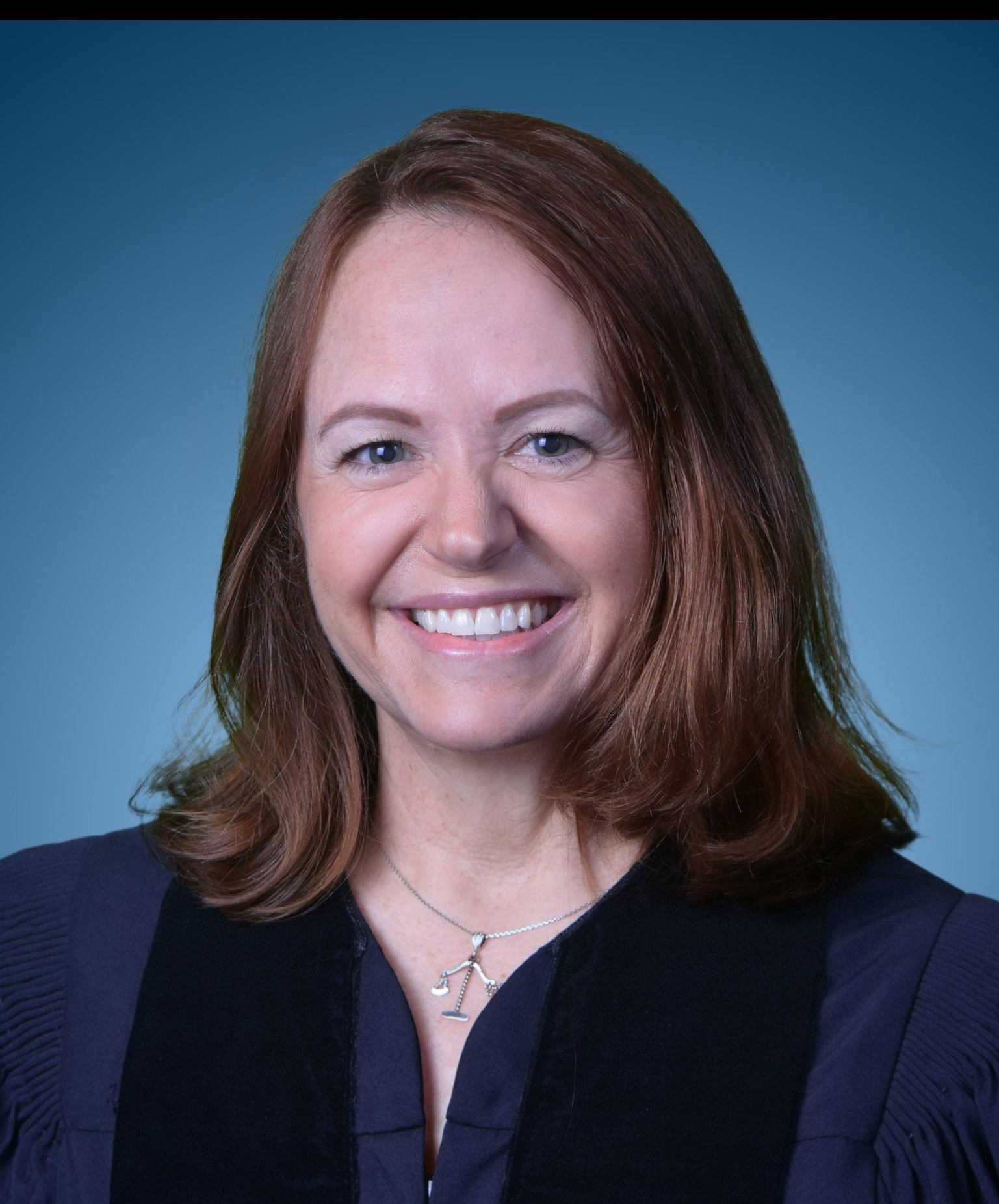 Headshot of a smiling woman with shoulder-length auburn hair wearing a dark blazer and necklace against a blue background.