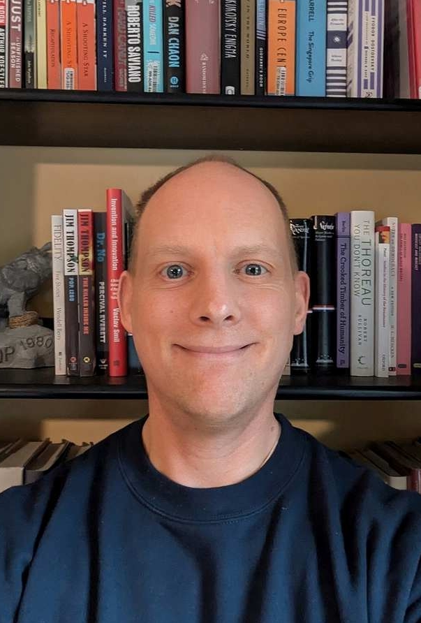 Smiling person taking a selfie in front of a bookshelf filled with books.