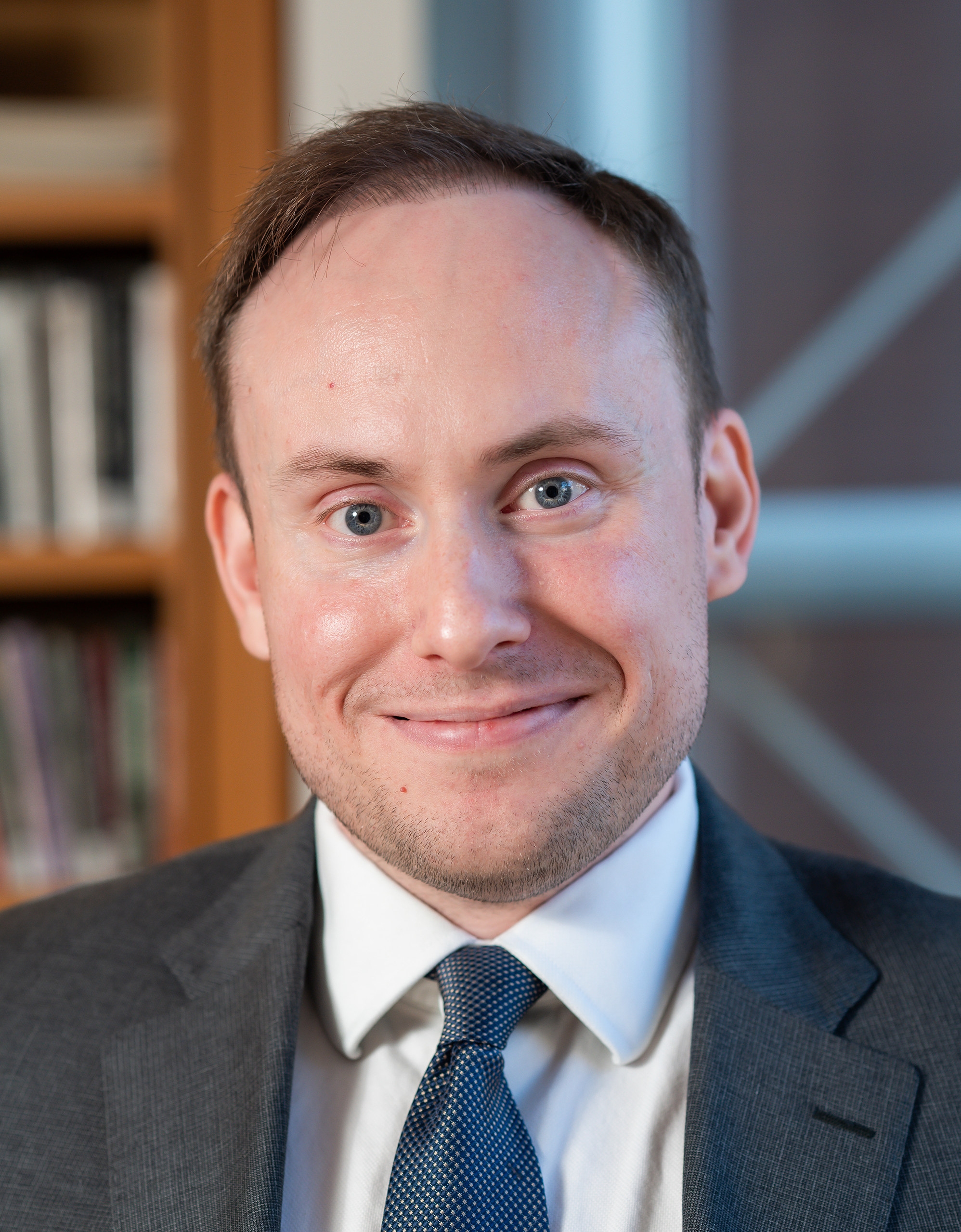 Close-up headshot of a smiling man in a suit and tie, with bookshelves blurred in the background.