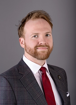 Studio headshot of a bearded man in a gray plaid suit with a red tie against a gray background.