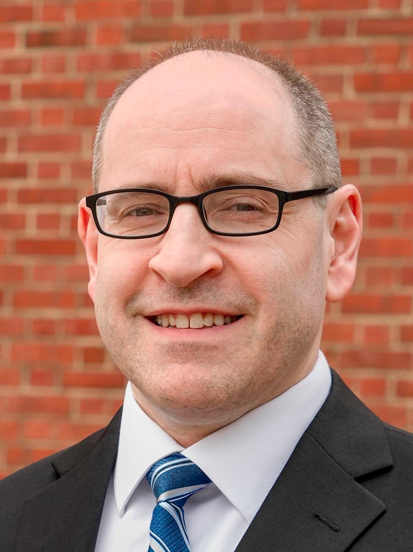 Close-up headshot of a smiling man wearing black rectangular glasses, a dark suit, and a blue striped tie, with a brick wall in the background.