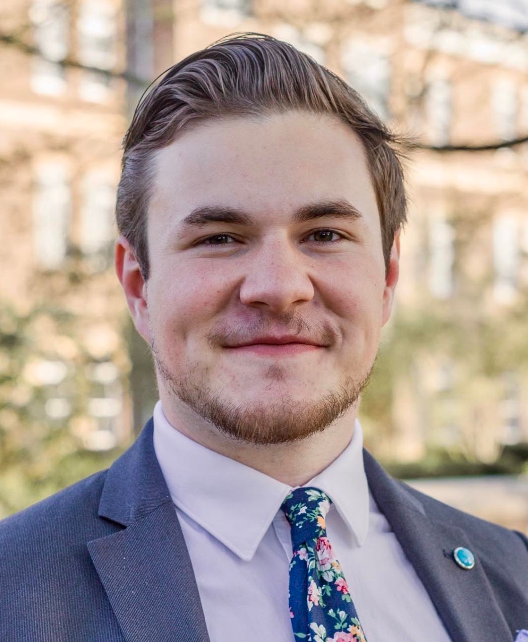 Close-up outdoor headshot of a smiling man in a suit and floral tie, with a softly blurred building and trees in the background.