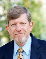 Head-and-shoulders portrait of a middle-aged man with short brown hair and a gray goatee, wearing a navy suit, light blue shirt, and patterned tie, outdoors with a blurred green background.