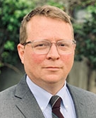 Portrait of a man wearing glasses and a gray suit with a striped tie, standing outdoors with a blurred background.