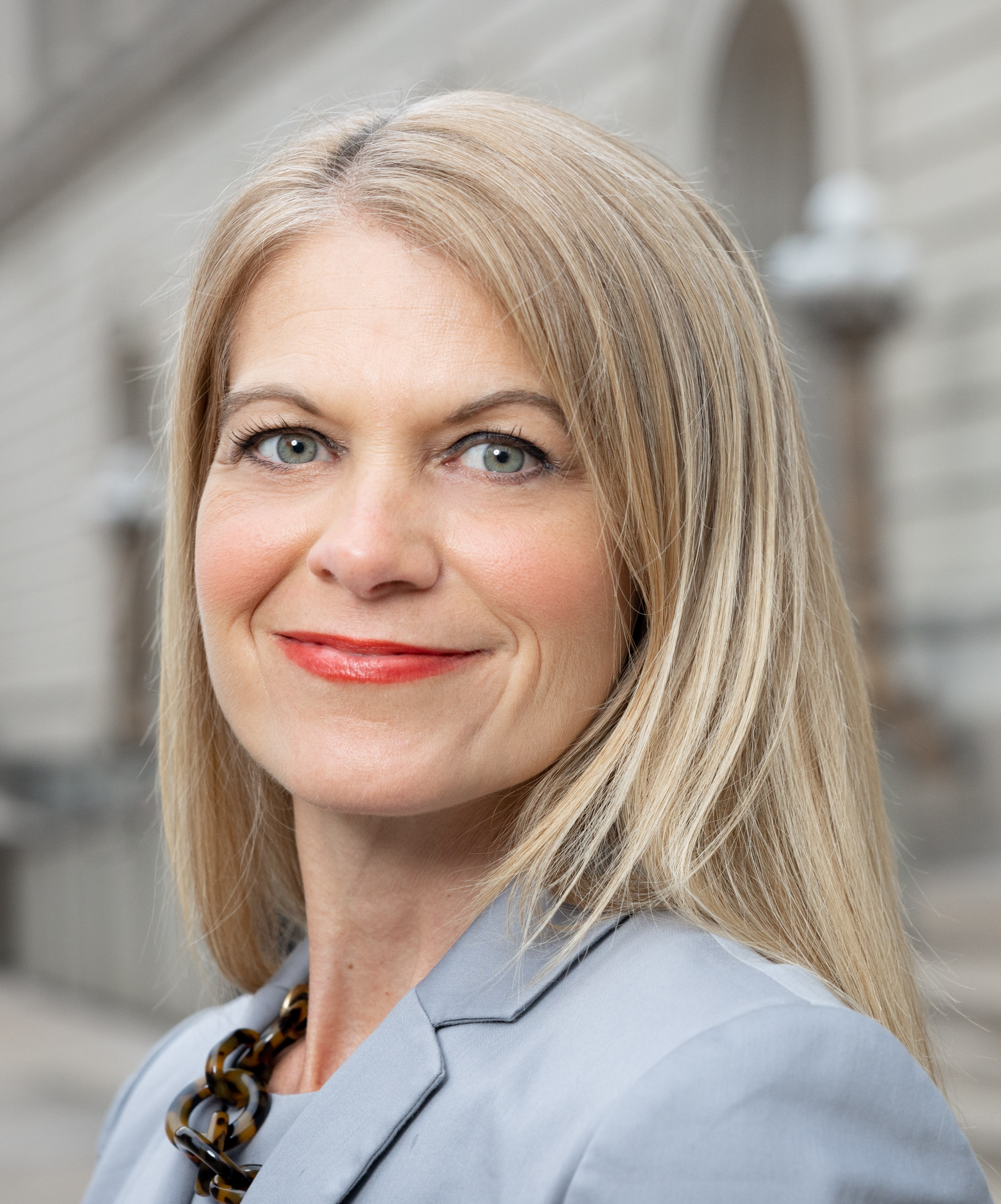 Close-up portrait of a blonde woman with red lipstick wearing a light gray jacket, against a softly blurred outdoor background.