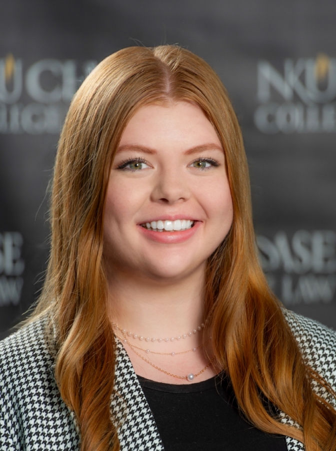 Head-and-shoulders portrait of a smiling woman with long red hair, wearing a patterned blazer and layered necklaces against a blurred event backdrop.