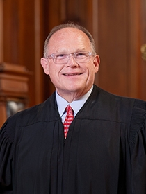 Smiling man wearing glasses and a black judicial robe with a red tie, standing indoors against a wood-paneled background.
