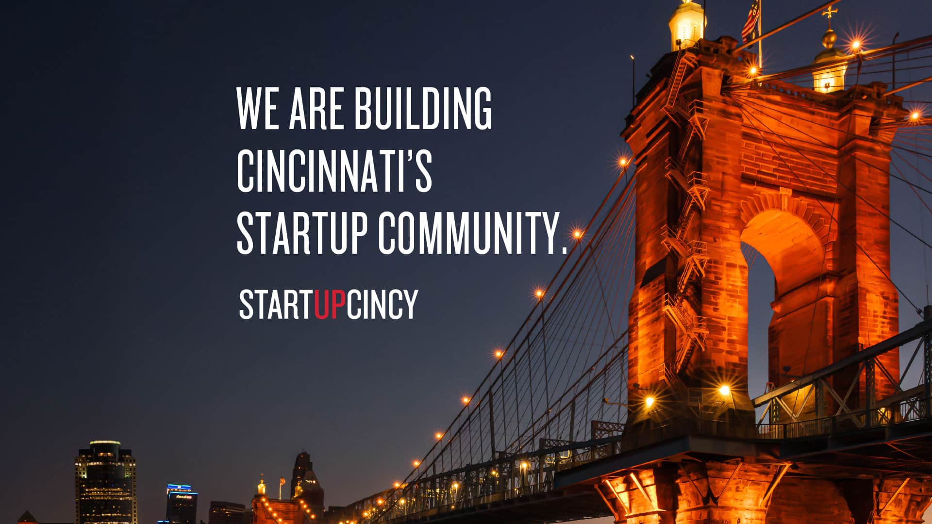 Night view of Cincinnati’s Roebling Suspension Bridge lit in orange, with the overlaid text “We are building Cincinnati’s startup community.” and “StartUpCincy.”.